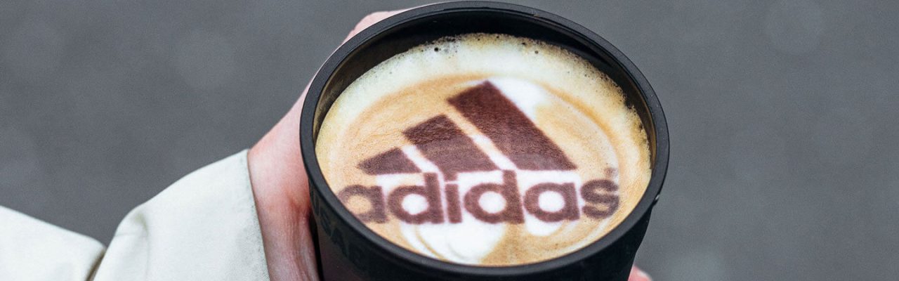 Kaffee in einem Reusable Cup. Im Kaffeeschaum sind das Logo und der Schriftzug von Adidas.