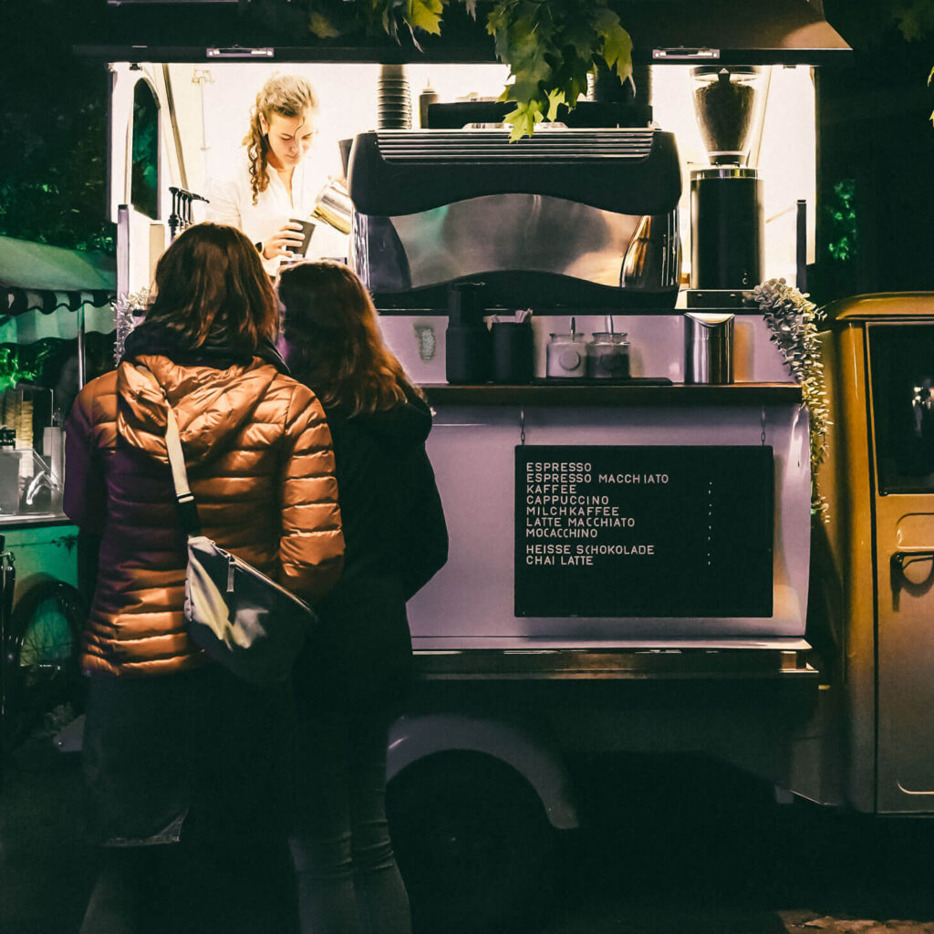Zwei Personen stehen vor der Kaffee-Ape bei Nacht. Die Barista in der Ape bereitet Kaffee zu.