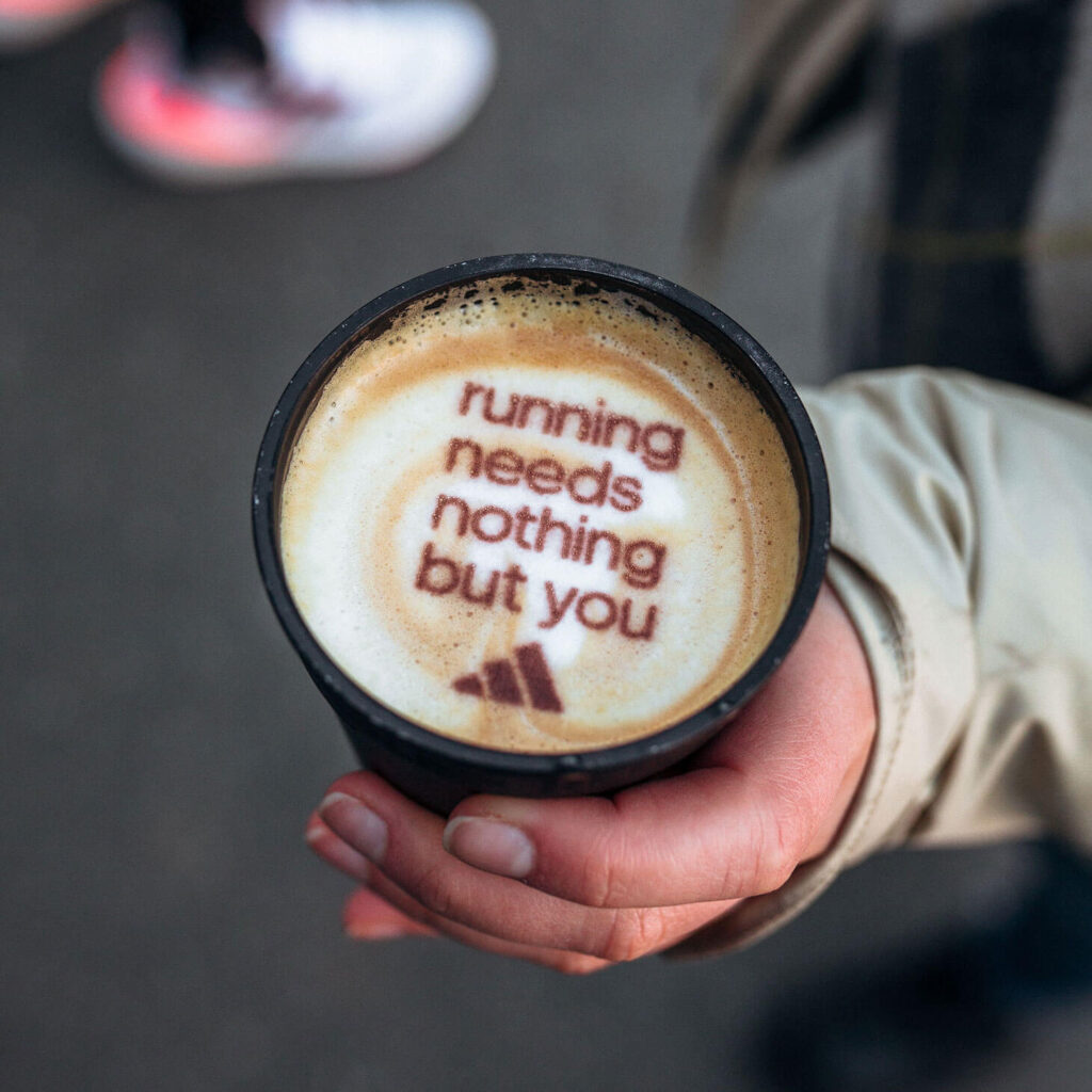 Eine Hand, in der ein Kaffeebecher gehalten wird. Auf dem Kaffeeschaum steht: running needs Nothing but you. Darunter ist das Logo von Adidas gedruckt.